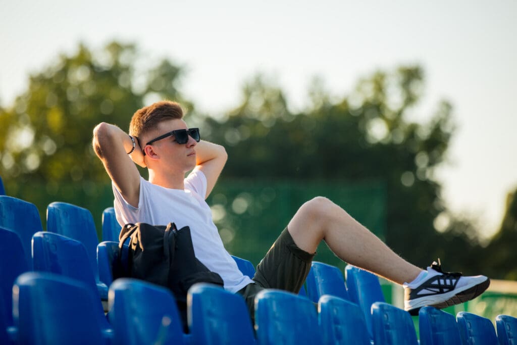 Young teen boy in sunglasses sitting in blue sit on stadium tribune